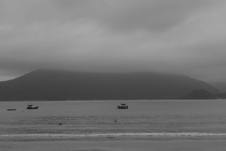 Vista em preto e branco do mar tranquilo de Ubatuba, com barcos e montanhas ao fundo.