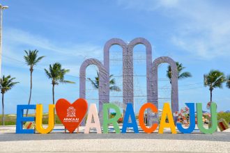 Letreiro ‘EU AMO ARACAJU’ diante de palmeiras, sob céu azul e ensolarado.