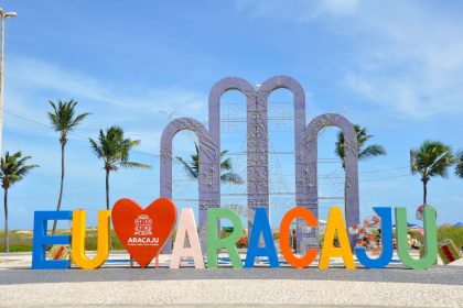 Letreiro ‘EU AMO ARACAJU’ diante de palmeiras, sob céu azul e ensolarado.
