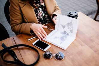 Pessoa consulta um planejador de viagem e um mapa sobre uma mesa de madeira, ao lado de uma xícara de café e um smartphone.