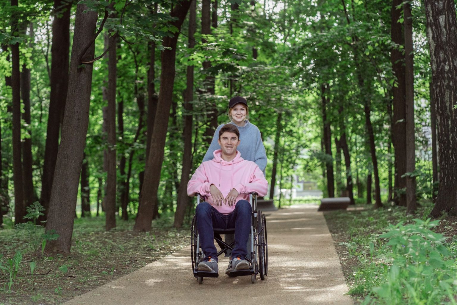 Casal em um parque ensolarado; uma mulher empurra um homem em cadeira de rodas por um caminho arborizado