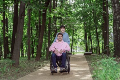 Casal em um parque ensolarado; uma mulher empurra um homem em cadeira de rodas por um caminho arborizado
